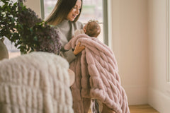 A mother holding her baby wrapped in the Hugs Heirloom Rose Blanket, showcasing its soft purple-pink hue.