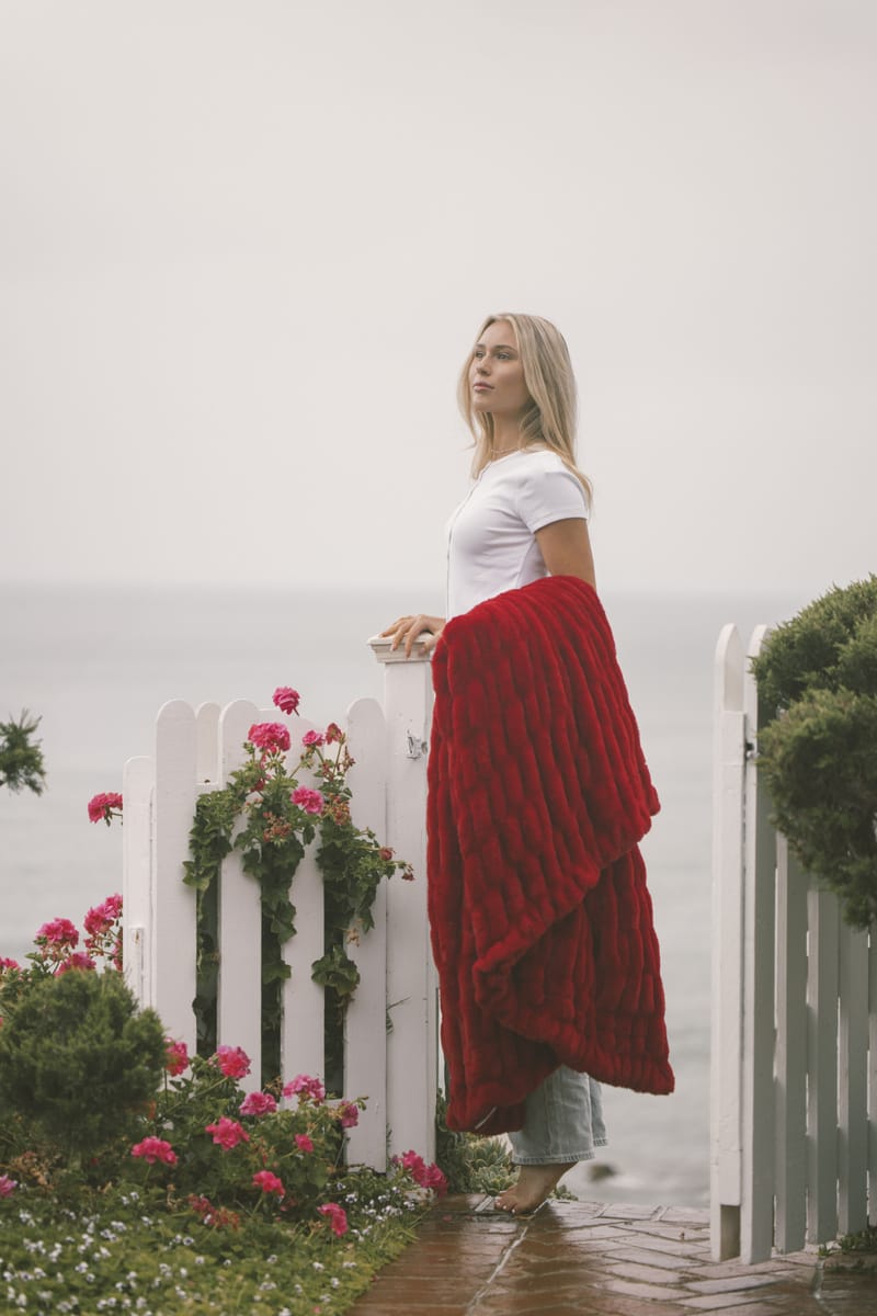Woman standing on a foggy path with a red Minky Couture blanket and white top, near a white picket fence with flowers.