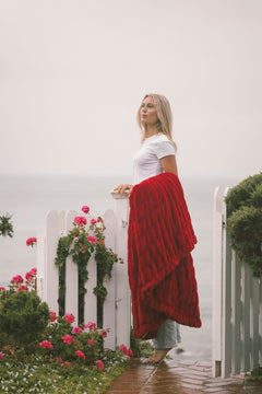 Woman standing on a foggy path with a red Minky Couture blanket and white top, near a white picket fence with flowers.
