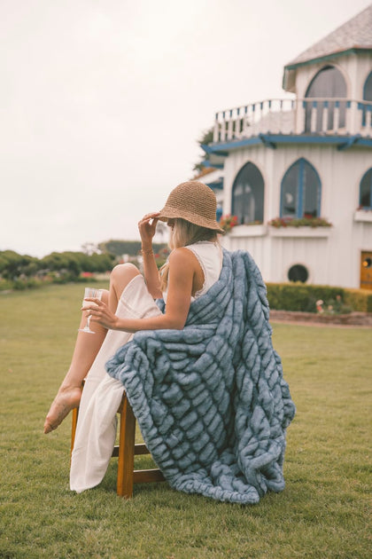Woman sitting on a chair with a blue Minky Couture Hugs blanket draped over her, holding a drink in a scenic outdoor setting.