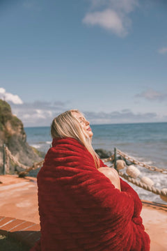 Person wrapped in a red Minky Couture blanket sitting by the ocean with a clear blue sky.