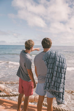 Two people standing by a railing overlooking the ocean with a clear sky with a blue Minky Couture blanket. 