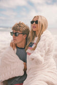 Two people wrapped in a cream Minky Couture Hugs blanket on a beach with sunglasses on