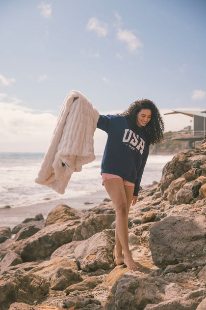 Person on rocks by the ocean wearing a navy blue 'USA' t-shirt holding a cream Minky Couture Hugs blanket.