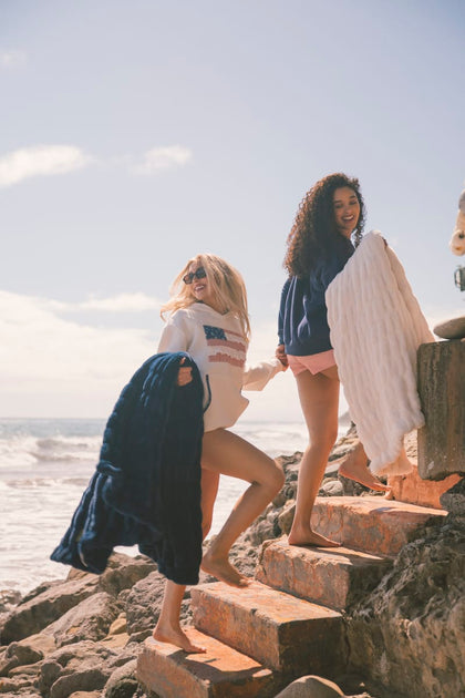 Two women standing on a rocky beach with ocean and sky in the background holding navy blue and cream Minky Couture Hugs blankets.