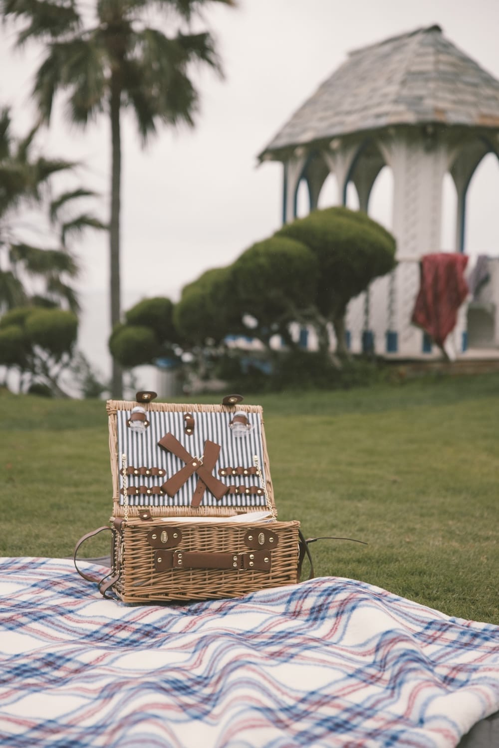 Wicker picnic basket on a plaid blanket with a gazebo and trees in the background