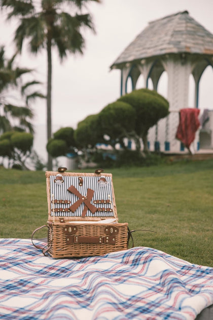 Wicker picnic basket on a plaid blanket with a gazebo and trees in the background