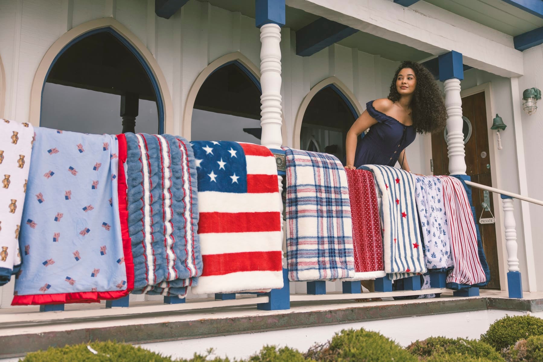 Woman standing on a porch with various patterned blankets, including an American flag design, draped over a railing.