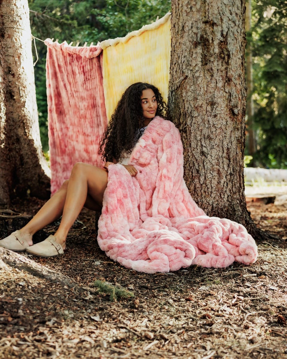 A woman sits on the forest floor between two trees, wrapped in a chunky pink Minky Couture blanket. A pink and yellow tie-dye blanket is hanging in the background among trees.