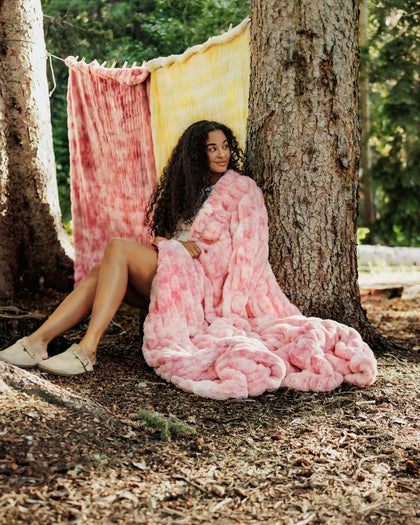 A woman sits on the forest floor between two trees, wrapped in a chunky pink Minky Couture blanket. A pink and yellow tie-dye blanket is hanging in the background among trees.