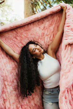 A woman with long curly dark hair lies on a fluffy Minky Couture Cotton Candy Pink blanket outdoors, holding the blanket open with both arms raised. She is wearing a white sleeveless top and light blue denim shorts.