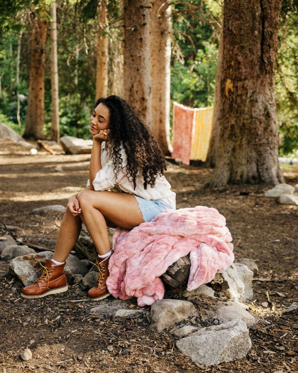 A woman sits on a stone in a wooded outdoor setting, wearing a white shirt, denim shorts, and brown lace-up boots, with a fluffy pink Minky Couture blanket draped over the stone beside her.