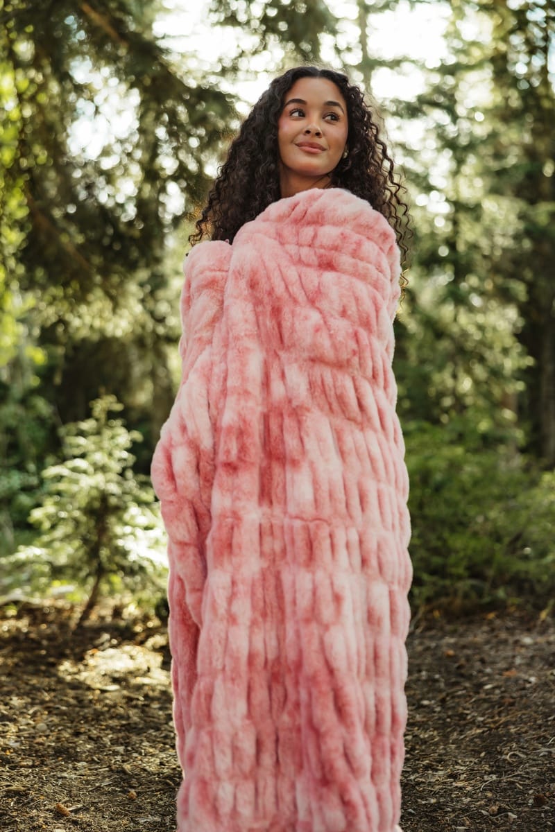 A woman wrapped in a chunky, textured pink Minky Couture blanket or wrap, standing in a sun-dappled forest setting.