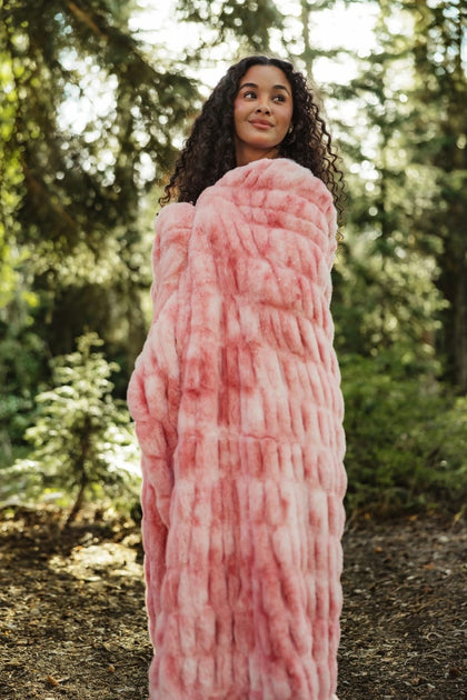 A woman wrapped in a chunky, textured pink Minky Couture blanket or wrap, standing in a sun-dappled forest setting.