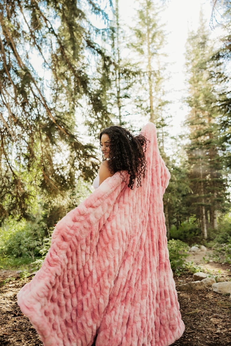 A woman wrapped in a chunky, fluffy pink Minky Couture blanket standing in a sunlit pine forest.