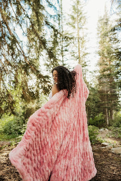 A woman wrapped in a chunky, fluffy pink Minky Couture blanket standing in a sunlit pine forest.