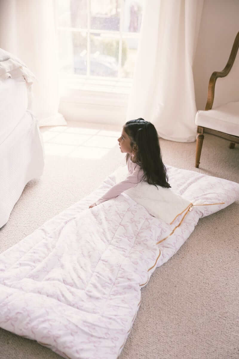 A girl sitting on the ground on a Minky Couture Sleeping Bag Pretty Pink Bows in a cozy room.