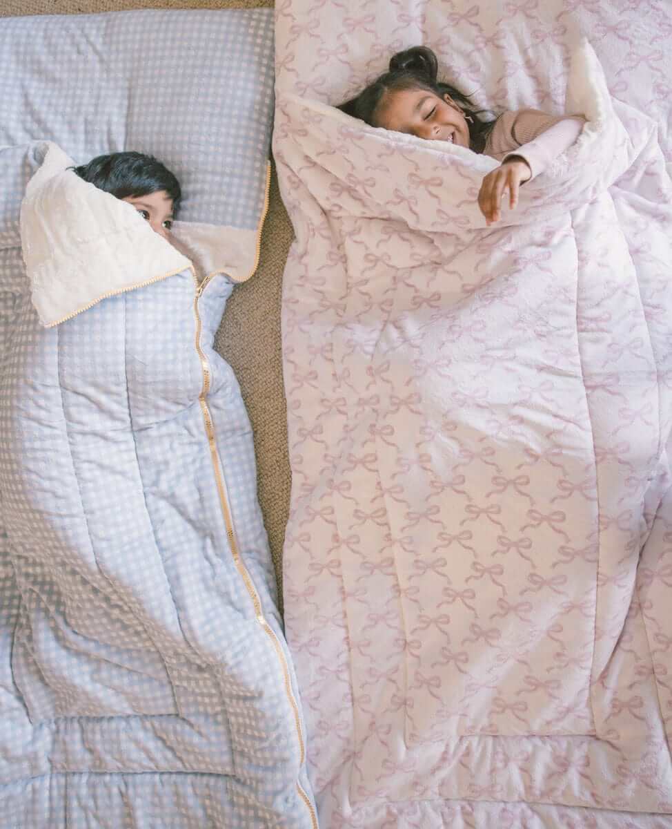 Two children nestled in Sleeping Bag Pretty Pink Bows and a blue patterned sleeping bag, enjoying a cozy moment.