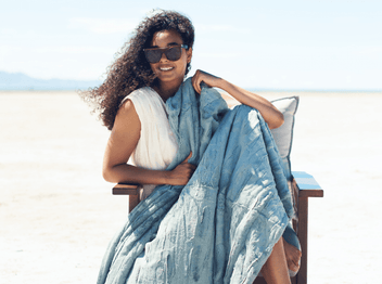 Woman in sunglasses draped in a blue blanket, sitting outdoors on a sunny day, exuding relaxed elegance.