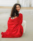 Woman sitting on the beach wrapped in a Sorbet Red Blanket for warmth and style.