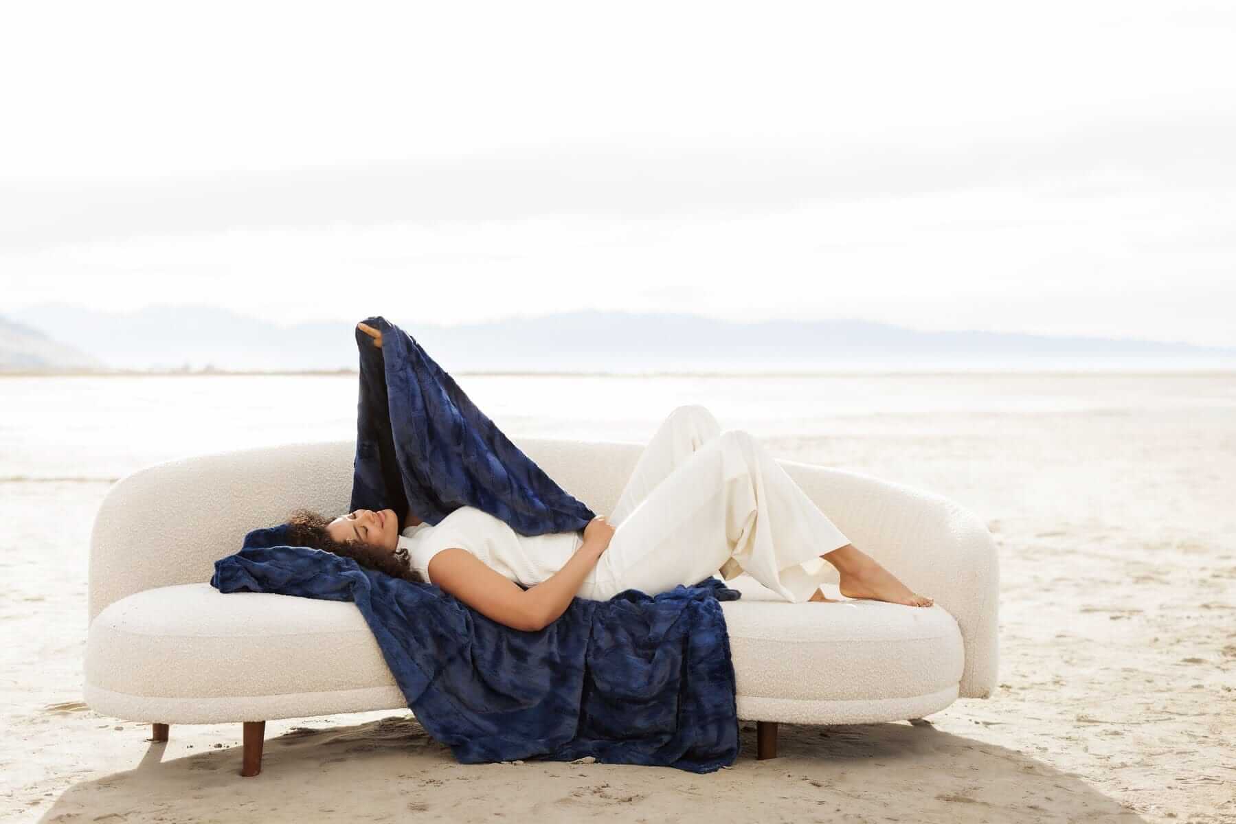 Woman relaxing on a couch draped with Sorbet Navy Blanket, showcasing its plush, textured finish and rich blue hue.