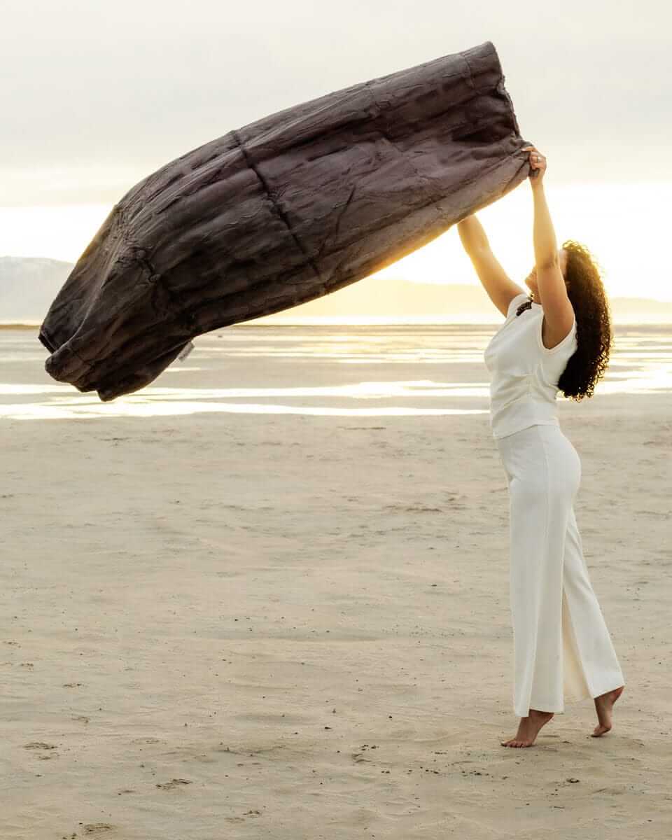 A woman joyfully holds the Sorbet Charcoal Blanket against a beautiful sunset backdrop on a sandy beach.
