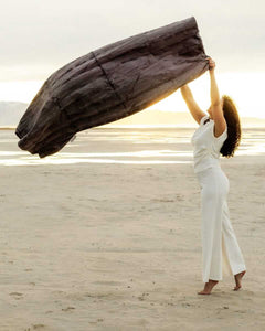 A woman joyfully holds the Sorbet Charcoal Blanket against a beautiful sunset backdrop on a sandy beach.