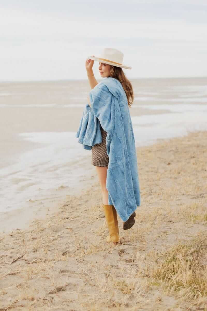 Model wearing a Sorbet Denim Blanket on a beach, showcasing its plush texture and soft blue hue.