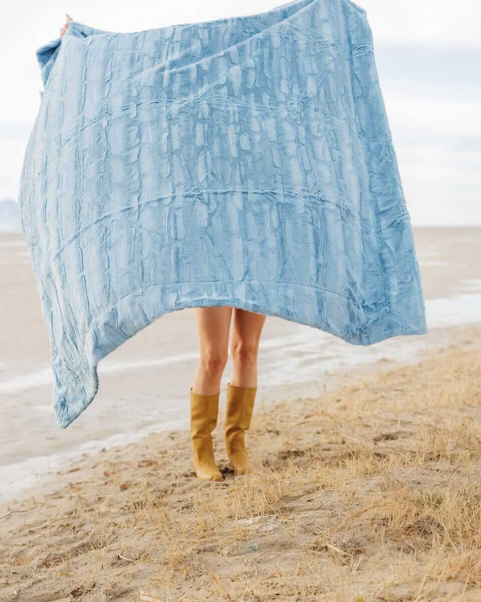 A person holding a Sorbet Denim Blanket in soft blue, showcasing its plush texture against a natural background.