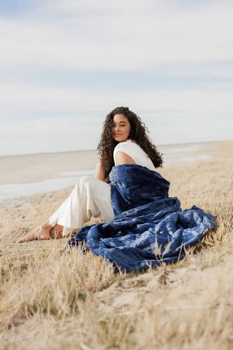 Woman sitting on the beach wrapped in a Sorbet Navy Blanket, showcasing its plush texture and comforting warmth.