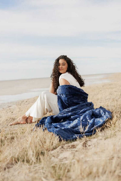 Woman sitting on the beach wrapped in a Sorbet Navy Blanket, showcasing its plush texture and comforting warmth.