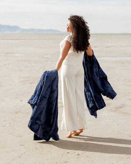 A woman walking on the beach holding two Sorbet Navy Blankets, showcasing their plush texture and rich blue hue.