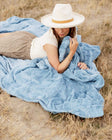 Woman relaxing on Sorbet Denim Blanket in muted blue, showcasing its plush texture and cozy appeal.