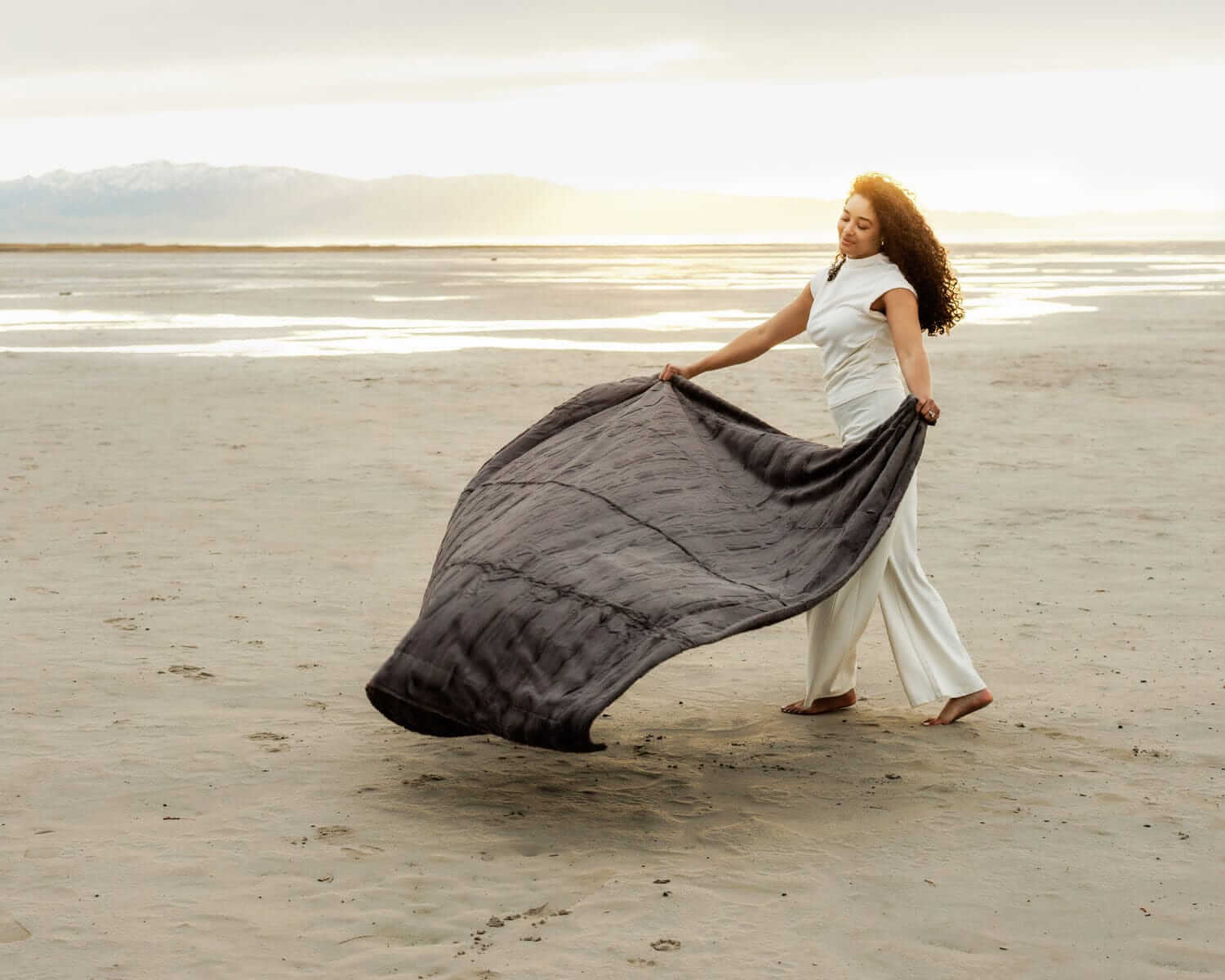 Woman elegantly drapes the Sorbet Charcoal Blanket on the beach at sunset, showcasing its plush texture.