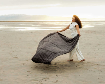 Woman elegantly drapes the Sorbet Charcoal Blanket on the beach at sunset, showcasing its plush texture.