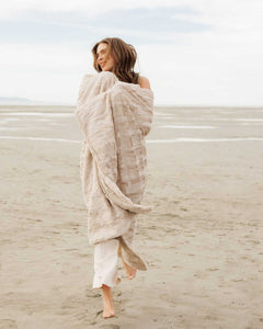 A woman wrapped in a soft Sorbet Almond Croissant Blanket on a sandy beach.