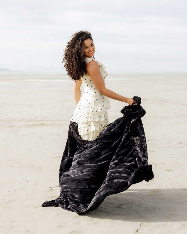 Woman in a white dress showcasing the luxurious Sorbet Black Blanket on the beach.