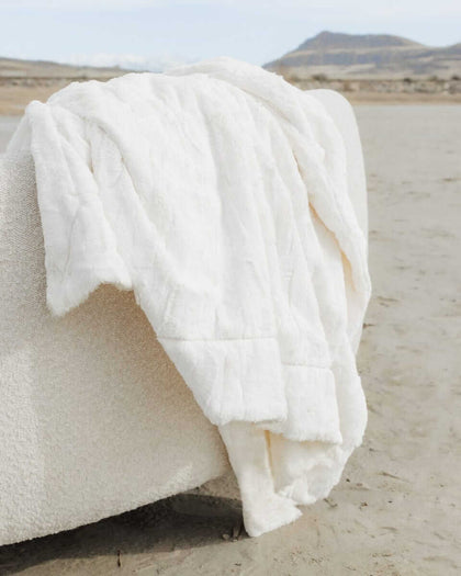 A Sorbet Vanilla Blanket draped over a chair on a sandy beach, showcasing its silky smooth cream texture.