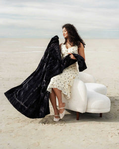 A woman in a polka dot dress happily holding the Sorbet Black Blanket on a beach, showcasing its elegant design.