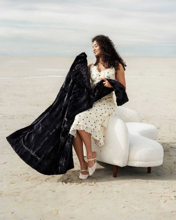 A woman in a polka dot dress happily holding the Sorbet Black Blanket on a beach, showcasing its elegant design.