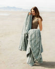 A woman holding the Sorbet Mist Blanket on a sandy landscape, showcasing its soft green tone and plush texture.