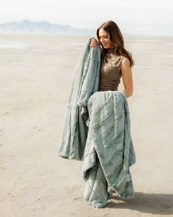 A woman holding the Sorbet Mist Blanket on a sandy landscape, showcasing its soft green tone and plush texture.