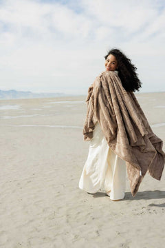 Woman wrapped in the Sorbet Milk Chocolate Blanket on a sandy beach, showcasing its plush, chocolatey texture.