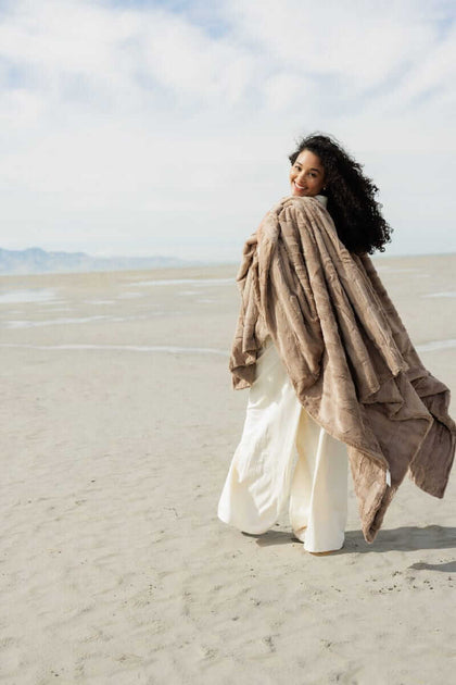 Woman wrapped in the Sorbet Milk Chocolate Blanket on a sandy beach, showcasing its plush, chocolatey texture.
