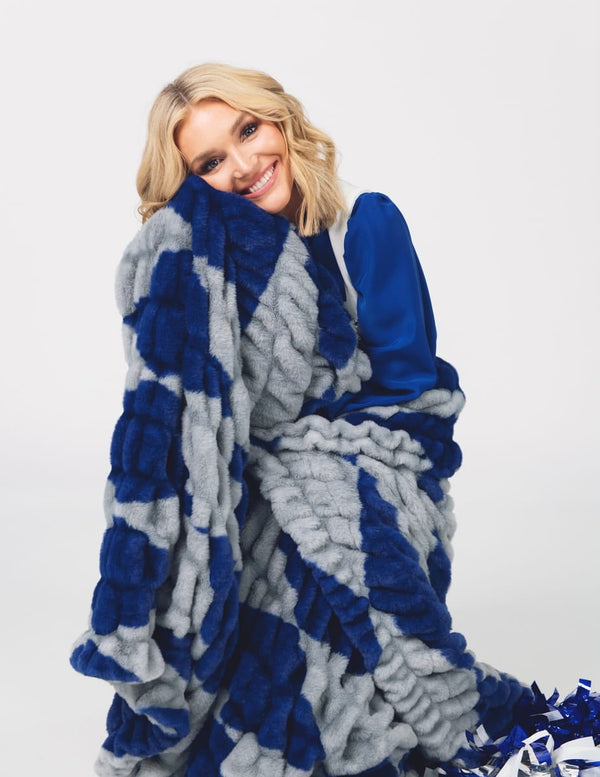 A smiling blonde woman wrapped in a chunky plush throw blanket with a bold blue and light gray checkered pattern, posed against a clean white studio background.