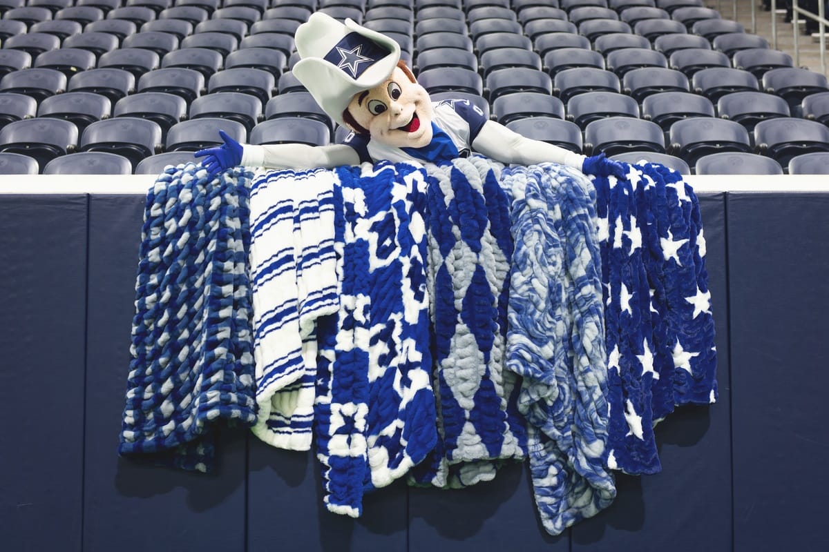 A Dallas Cowboys mascot character wearing a hat leans over a barrier draped with several blue and white patterned blankets, with rows of empty stadium seats in the background.