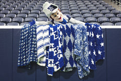 A Dallas Cowboys mascot character wearing a hat leans over a barrier draped with several blue and white patterned blankets, with rows of empty stadium seats in the background.