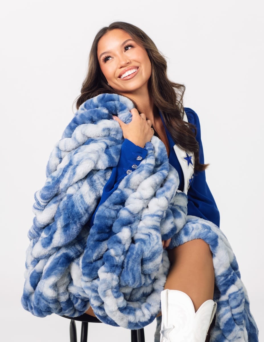 A smiling woman seated on a stool wrapped in a chunky, oversized blue and white marled throw blanket.
