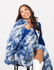 A smiling woman seated on a stool wrapped in a chunky, oversized blue and white marled throw blanket.