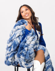 A smiling woman seated on a stool wrapped in a chunky, oversized blue and white marled throw blanket.